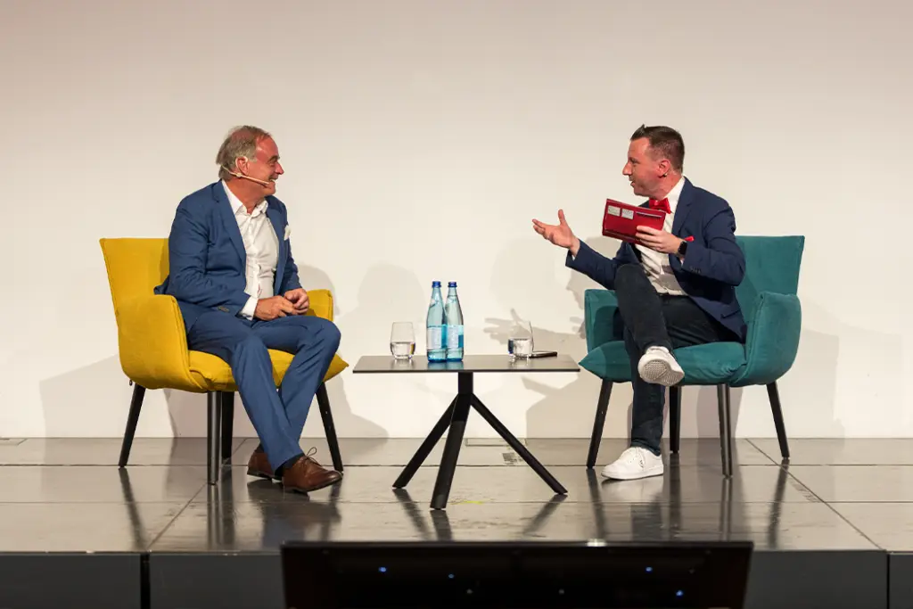 Torsten Schröder sitzt auf der Bühne auf einem Sessel, daneben ein Mann ebenfalls in einem Sessel, beide unterhalten sich