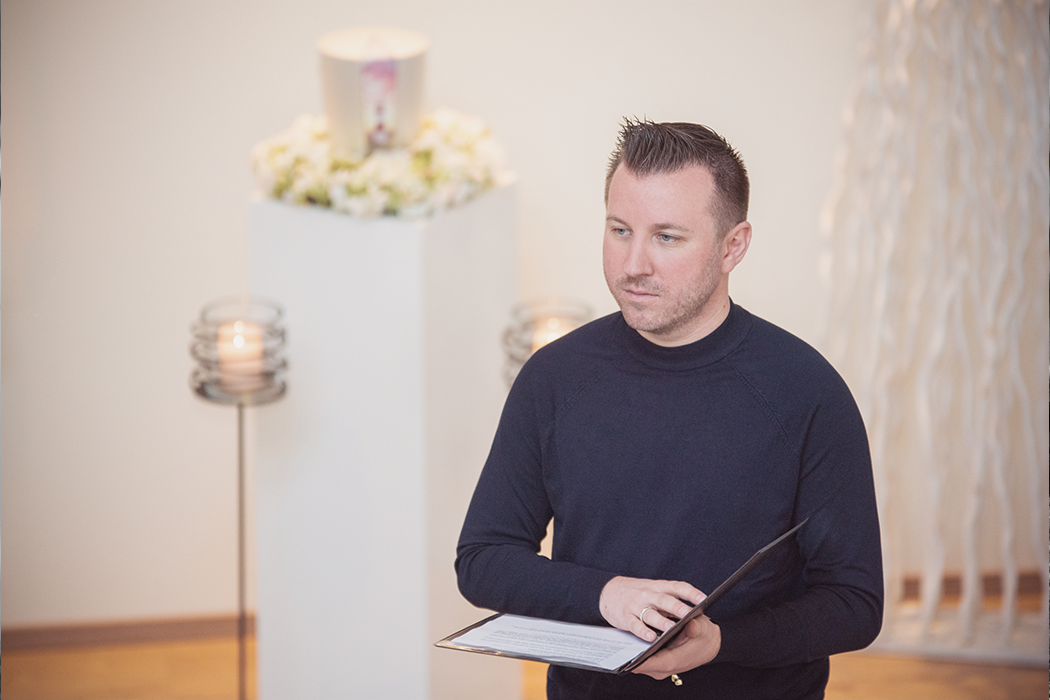 Torsten Schröder steht mit einem Buch in der Hand und ernster Mine da, im Hintergrund ist eine Urne zu erkennen